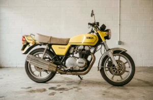 Suzuki GT750 jaune candy d'origine garée dans un garage vintage, vue latérale complète révélant le moteur deux-temps triple et les échappements chromés d'époque