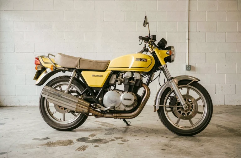 Suzuki GT750 jaune candy d'origine garée dans un garage vintage, vue latérale complète révélant le moteur deux-temps triple et les échappements chromés d'époque
